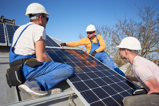 Male team workers installing stand-alone solar photovoltaic panel system. Electricians mounting blue solar module on roof of modern house. Alternative energy ecological concept.