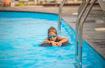 Naklejka premium Charming little girl in swimming goggles and a lifebuoy swims in the clear blue water of the pool during summer vacation. Concept of children's holidays