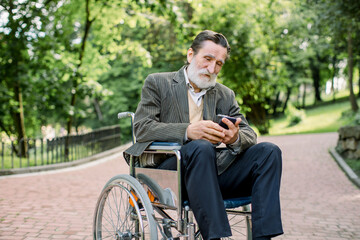 Bearded Caucasian old man is sitting in a wheelchair in the park alone, using his smartphone for serfing internet or socail networks