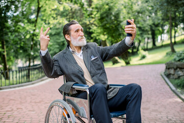 Happy senior disabled or handicapped bearded man sitting on a wheelchair in park, using his smartphone for photographing himself on the background of park landscape
