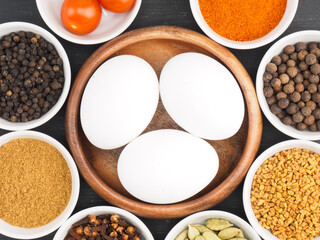 Raw chicken eggs, black pepper, masala powder, fenugreek, green cardamom, hot red chili pepper, cloves, tomato on black wooden background. Ingredients for making scrambled eggs or omelet. 