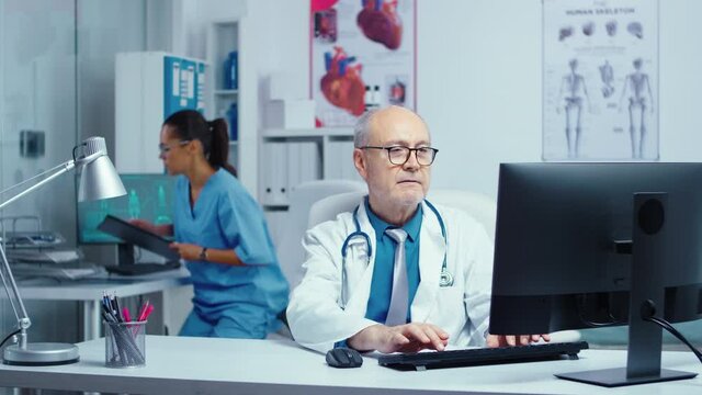 Senior doctor in moden private clinic working at PC then smiling at the camera. Healthcare specialist medicine practitioner in uniform working in hospital with big staff
