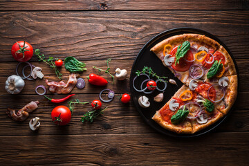 Fototapeta na wymiar Pizza with bacon and ham without one slice on a slate black platter which is on wooden background, top view and copy space