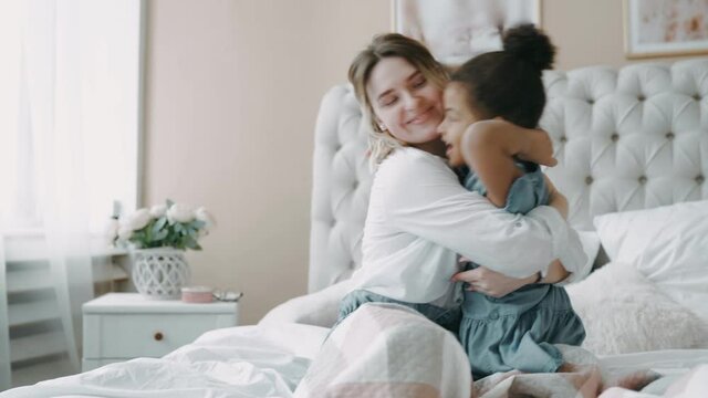Happy loving young mother european in white shirt with her charming little african daughter on big light bed in bedroom in morning. Caring mom helped button up baby blue dress, hugging and kissing her