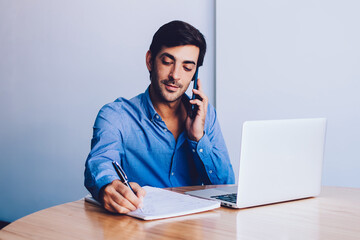 Serious entrepreneur planning work process writing main notes in textbook while talking with colleague about business deal using mobile phone for conversation sitting at table with laptop computer