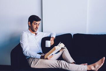 Young hipster guy enjoying literature hobby resting with book on leisure during morning coffee, handsome indian man relaxing on comfortable sofa reading best seller and drinking caffeine beverage