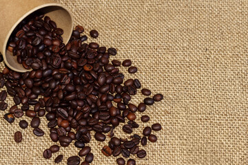 Roasted coffee beans spills out from a brown paper cup on sackcloth background.