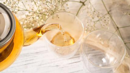 From transparent glass teapot pour golden tea in glass mug. Glass teapot pouring black tea into cup. White wooden table