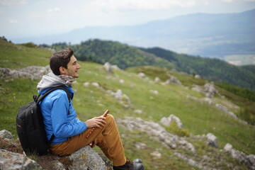 A young male tourist sitting on a rock. He is on the summit of Stara Planina (Balkan Mountains) and enjoying the view. In one of his hands, he's holding a wooden flute/ pipe.
