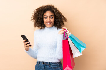 Young African American woman isolated on beige background holding shopping bags and a mobile phone