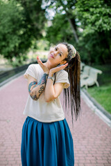 Naklejka premium Creative hipster girl with dreadlocks and tattoos in a city park on sunny summer day. Young stylish girl with dreadlocks outdoors