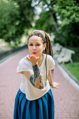 Front view of beautiful young hipster woman with dreadlocks hair style and tattoo, strolling in summer park, posing to camera and making air kiss