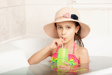 Funny little caucasian beautiful girl in hat plays beach bathing at home in bathroom and drinks cocktail. Concept of relaxation at sea. Entertainment at home during quarantine