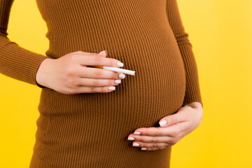 Close up of pregnant woman in brown dress with a cigarette against her belly at yellow background. Dangerous risk for unborn baby