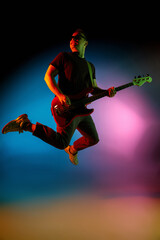 Jumping high. Young inspired and expressive musician, guitarist performing on gradient colored background in neon. Concept of music, hobby, festival, art. Joyful artist, colorful, bright portrait.