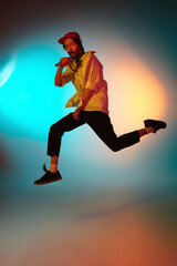 Fototapeta na wymiar Jumping high. Young inspired and expressive musician, singer performing on gradient colored background in neon light. Concept of music, hobby, festival, art. Joyful artist, colorful, bright portrait.