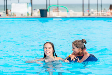 Fototapeta premium Two little sisters girls are swimming in a large pool with clear blue water of mine near the hotel on the background of the sea and the beach. Concept vacation tropical hot country with children