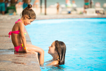 Fototapeta na wymiar Side view little girls sisters swim in the pool and splashes water on each other during a vacation in a tropical country on a sunny warm summer day. Children rest concept