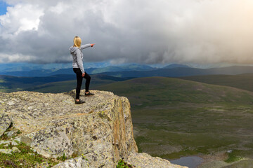 Obraz premium Young blonde girl in gray sweatshirt with black leggings stands on a cliff in the Altai mountains and points outstretched finger to the horizon where there is a picturesque landscape with many hills.