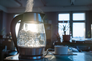 A transparent kettle of water boils against the background of the sunset shining through the...