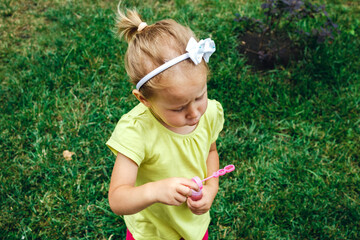 Summer outdoor activities for children. A little blonde girl in a yellow t-shirt blows soap bubbles against the background of green bright grass in the courtyard with a blur of focus.