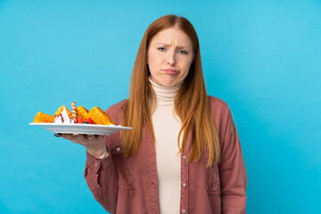 Young redhead woman holding waffles over isolated background with sad expression