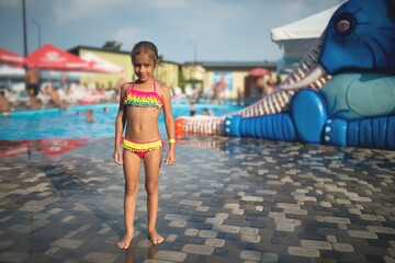 Little slim cute girl in bright swimsuit poses on backdrop of children's water zone in an open-air on warm summer day during vacation. Concept of children's entertainment and outdoor activities.