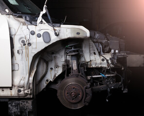 The front of the car after an accident with a disassembled body and metal elements with the wheel removed on a rack under the hood in a car service or garage against a dark background.