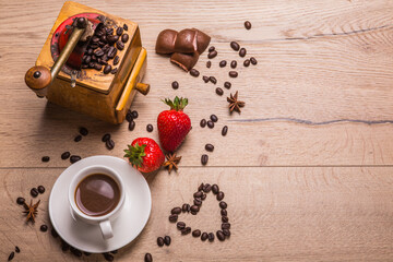 White cup of coffee stands on wooden table. Nearby there is vintage coffee grinder with hand drive, scattered coffee beans, heart, anise stars, two ripe strawberries, airy chocolate.