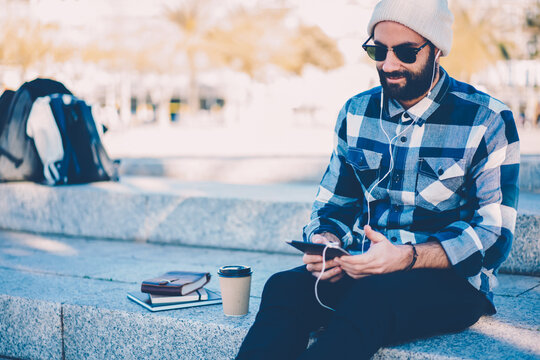 Bearded casual dressed hipster guy in sunglasses and hat listening online course about learning foreigh languages in modern earphones connected to modern touch pad device sitting in urban setting