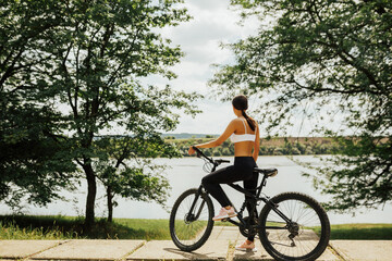Fototapeta premium Beautiful warm summer day. Back portrait of young attractive brunette woman in sportswear enjoying beautiful landscape after bicycle ride. Park. Sunny summer day.