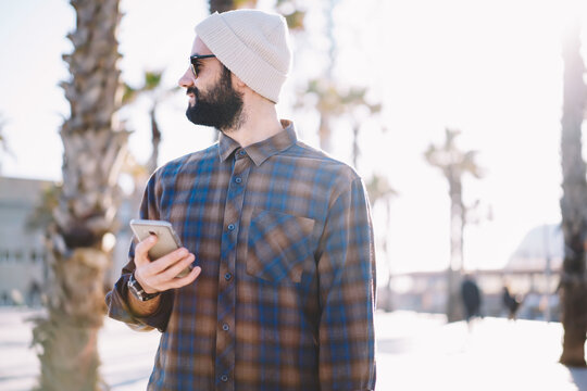 Handsome bearded male tourist strolling during sitty touch searching right direction via application with online maps on smartphone,hipster guy in sunglasses and hat standing on promotional background