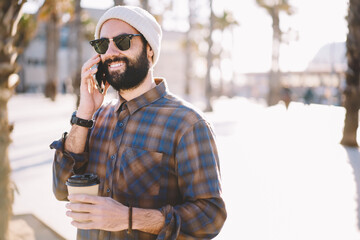 Smiling bearded tourist in hat and sunglasses strolling during city tour communicating with friends in roaming, handsome hipster guy having phone conversation standing near copy space for advertising