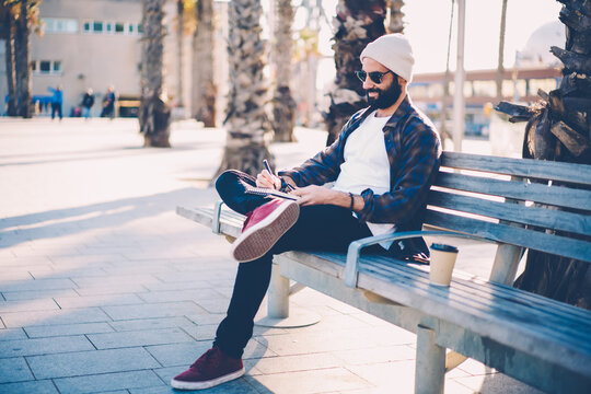 Handsome talented male painter resting on wooden bench drawing scketches in notepad about travelling enjoying working outdoors in sunny day.Smiling bearded guy making notes of his trip in notebook