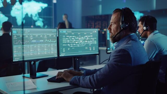 Confident Male Data Scientist Works on Personal Computer Wearing a Headset in Big Infrastructure Control and Monitoring Room. Senior Engineer in a Call Center Office Room with Colleagues.