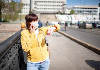 Funny middle-aged woman in a white protective mask talking on a smartphone while walking around the city on a warm spring day. Coronovirus pandemic and remote communication concept