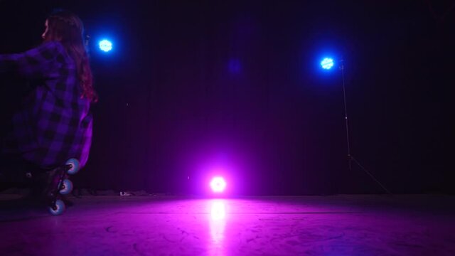 Professional roller skater girl crouches on rear and front wheels in scene, approaching view of pink spotlight between blue lamps. Young woman rides past on roller skates. Performance on rollerblades.
