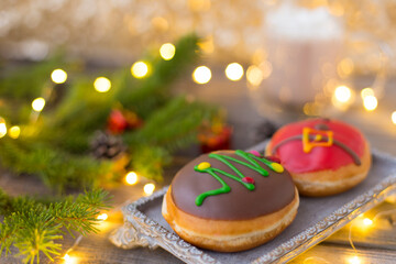 Christmas chocolate donuts with Santa Claus icing and new year tree on ceramic stand. Glass cup of cocoa with marshmallows on wooden table. Spruce branch, present, cone, sugar bowl, tea pot, garland