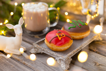 Christmas chocolate donuts with Santa Claus icing and new year tree on ceramic stand. Glass cup of cocoa with marshmallows on wooden table. Spruce branch, present, cone, sugar bowl,white deer,garland