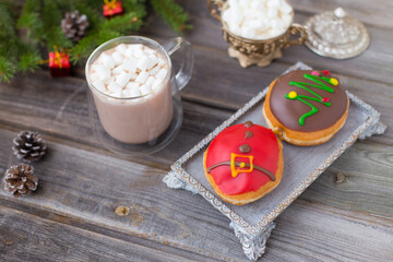 Christmas chocolate donuts with Santa Claus icing and new year tree on ceramic stand. Glass cup of cocoa with marshmallows on wooden table. Spruce branch, present, cones, sugar bowl, white deer