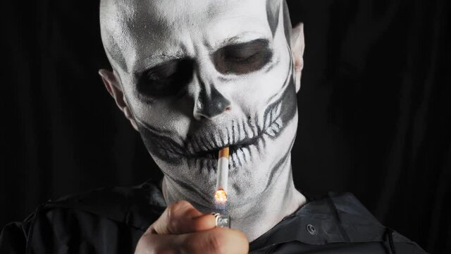 Man with make-up skeleton and black hood on a dark background. Skeleton smokes a cigarette. Smoking kills. Halloween or horror theme. High quality 4k video