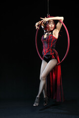 A adul girl in red evening dress performs the acrobatic elements in the air ring on a black background.