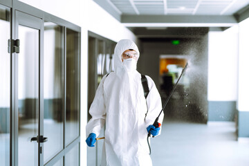 Fototapeta premium Contractor in a protective suit and mask sprays disinfectants in office before work. Protection against COVID-19 disease. Prevention of spreding pneumonia virus with surfaces.