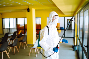 Obraz premium Contractor in a protective suit and mask sprays disinfectants in office before work. Protection against COVID-19 disease. Prevention of spreding pneumonia virus with surfaces.