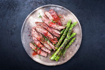 Traditional barbecue sliced dry aged wagyu flank steak offered with green asparagus and chili powder offered as overhead view on a rustic modern design plate with copy space
