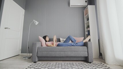 Young attractive Caucasian woman tapping, scrolling and typing on the smartphone while lying on the sofa at home