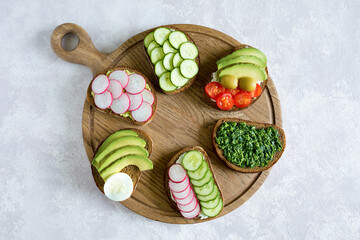 Vegetarian sandwiches on the wood board on the gray background. Fresh bread with pesto, cucumber, avocado, cherry tomatoes, radish and cheese. Delicious breakfast. Flat lay, top view.