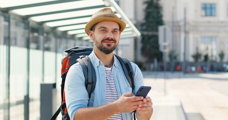 Caucasian young handsome man in hat with backpack tapping and scrolling on smartphone and smiling...
