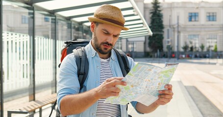 Young handsome Caucasian man tourist in hat with backpack holding city map and planning route....