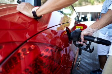 Cropped shot view of a young man pumping gasoline fuel in his luxury red car cabriolet at gas station while stopping during road trip with friends, transportation and expenses concept - obrazy, fototapety, plakaty